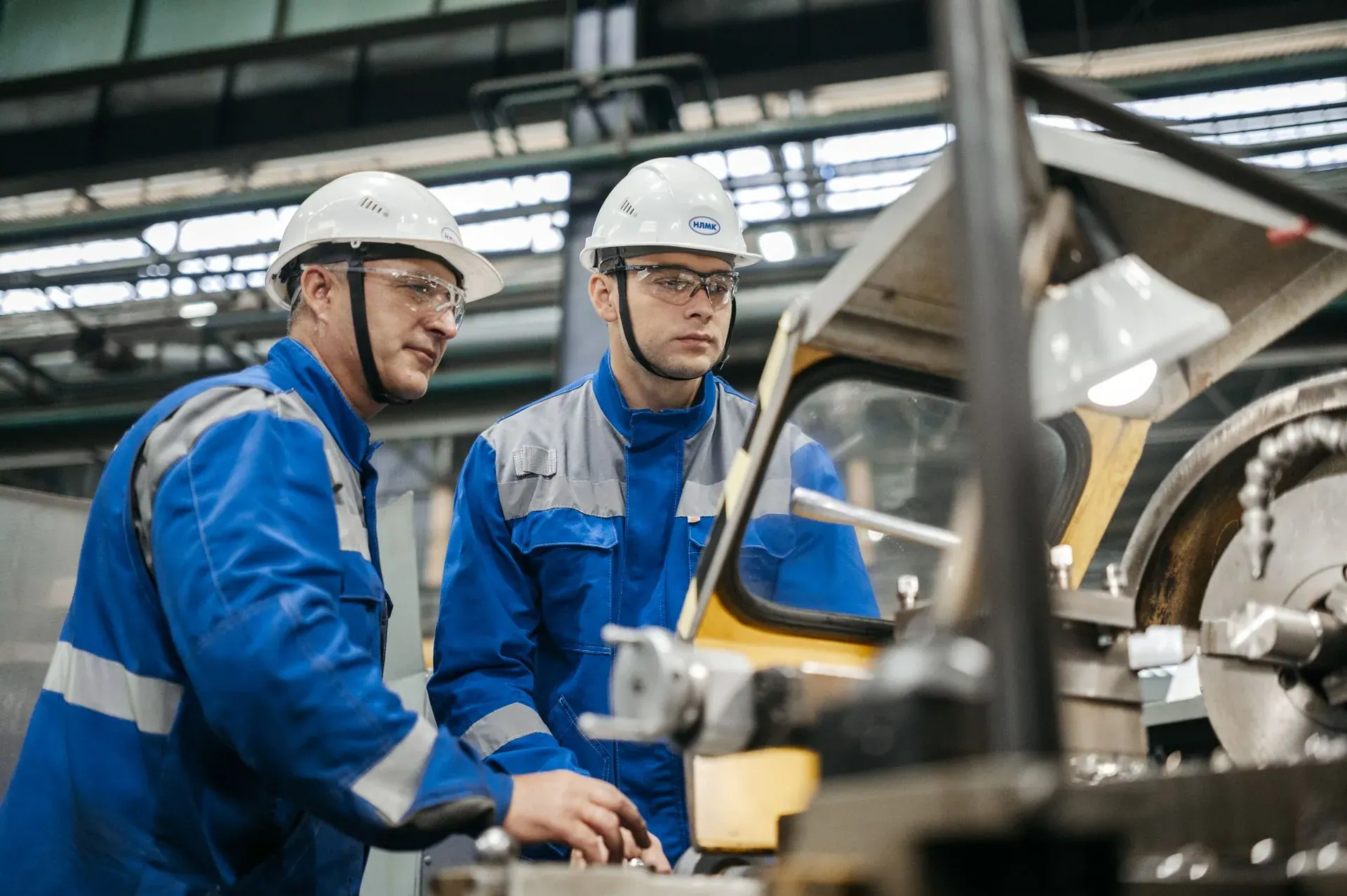 Two workers in overalls and hard hats operating a machine together on a factory floor